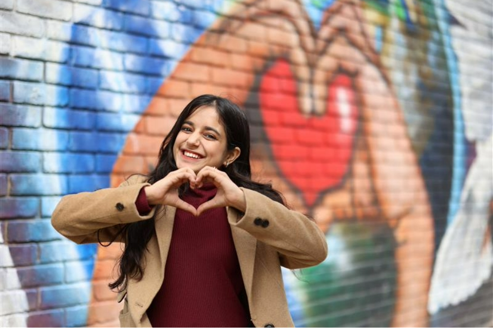 A young girl stans in front of a mural of hands holding a heart. She is making a heart symbol with her hands.